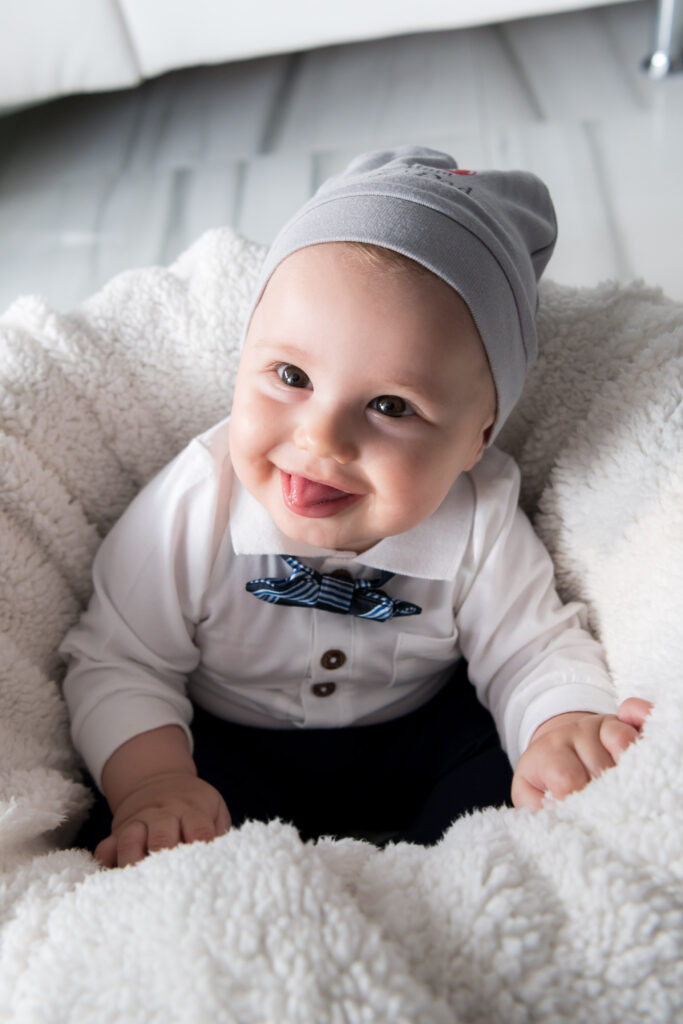 Lachendes Baby mit grauer Mütze und Fliege auf flauschiger Decke streckt die Zunge heraus