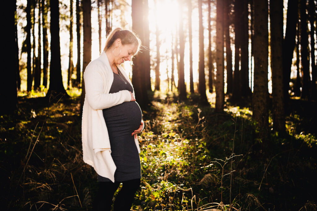 Schwangere umfasst lächelnd ihren Babybauch im sonnendurchfluteten Wald, stimmungsvolles Babybauch-Shooting