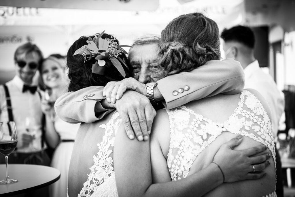 Älterer Mann umarmt gerührt zwei Bräute bei der Feier, emotionaler Schwarzweiß-Moment
