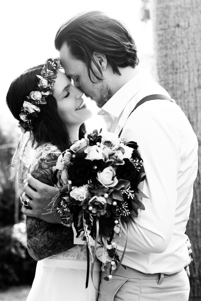 Boho-Brautpaar Stirn an Stirn, Braut mit Blumenkranz und Strauß, zärtlicher Moment in Schwarzweiß