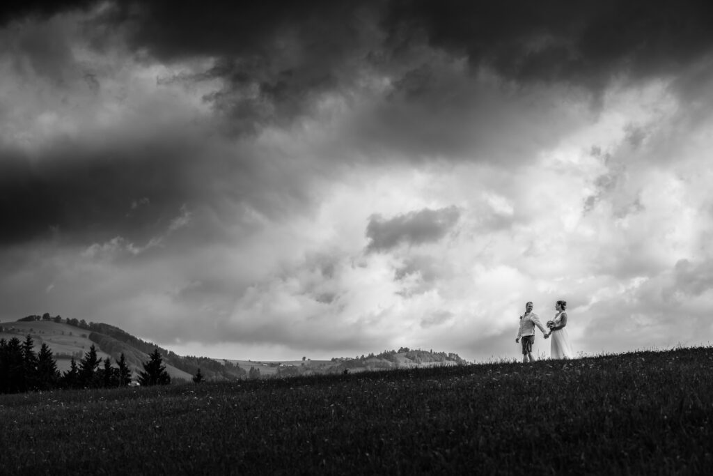 Brautpaar Hand in Hand auf Hügelkamm unter dramatischem Wolkenhimmel, Schwarzweiß