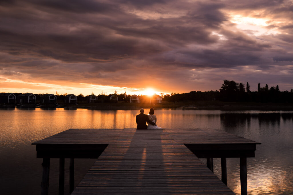 Brautpaar sitzt eng umschlungen auf einem Steg vor dramatischem Sonnenuntergang am See