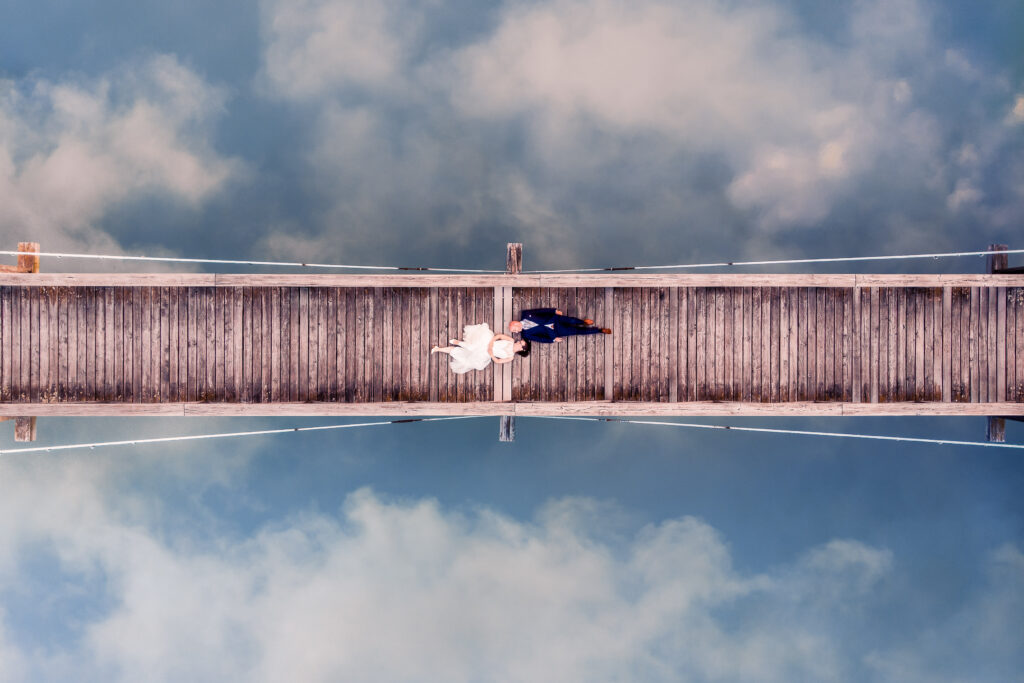 Vogelperspektive auf Brautpaar Hand in Hand auf Holzsteg, Wolken spiegeln sich im Wasser