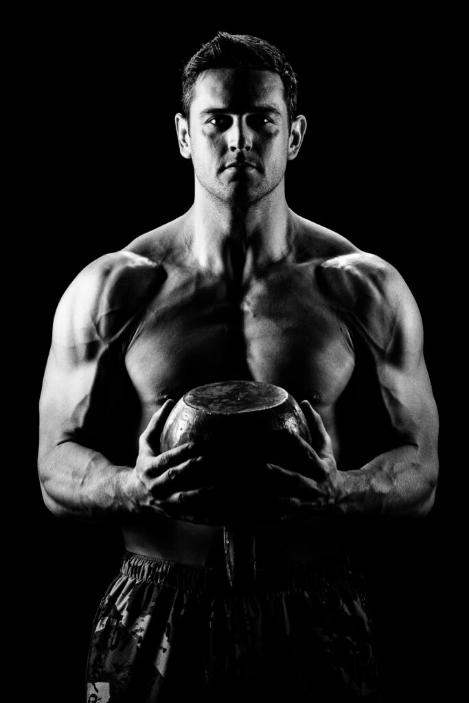Muskulöser Mann hält Kettlebell vor der Brust, kontrastreiches Schwarzweiß-Portrait vor schwarzem Hintergrund